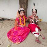 Two young girls in bright traditional indian costumes sit on sand beside a tree wearing ornate headpieces and jewelry one in pink with a flower crown and orange veil the other in red with a decorative crown holding a toy sword